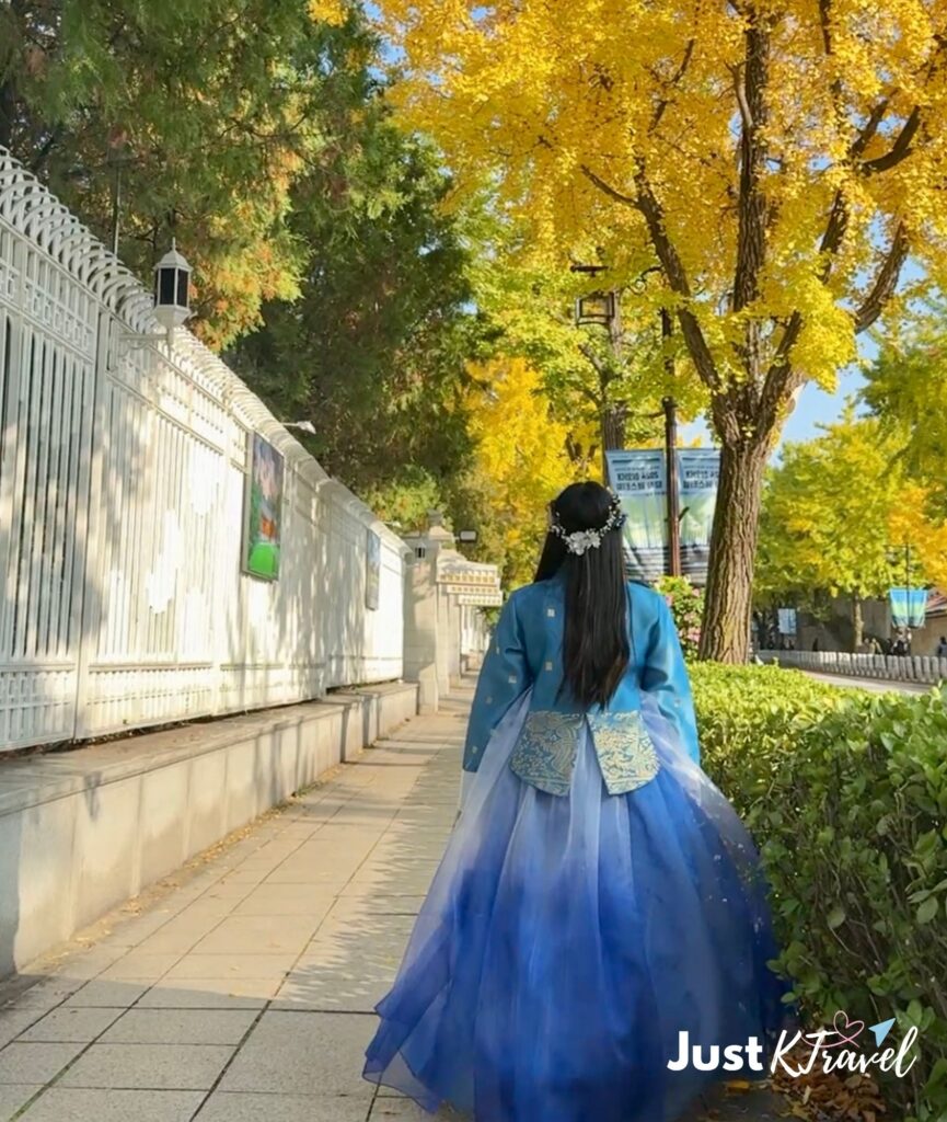 Person wearing traditional hanbok walking along Hyoja-ro (효자로) in Seoul during autumn, surrounded by colorful fall foliage and golden trees