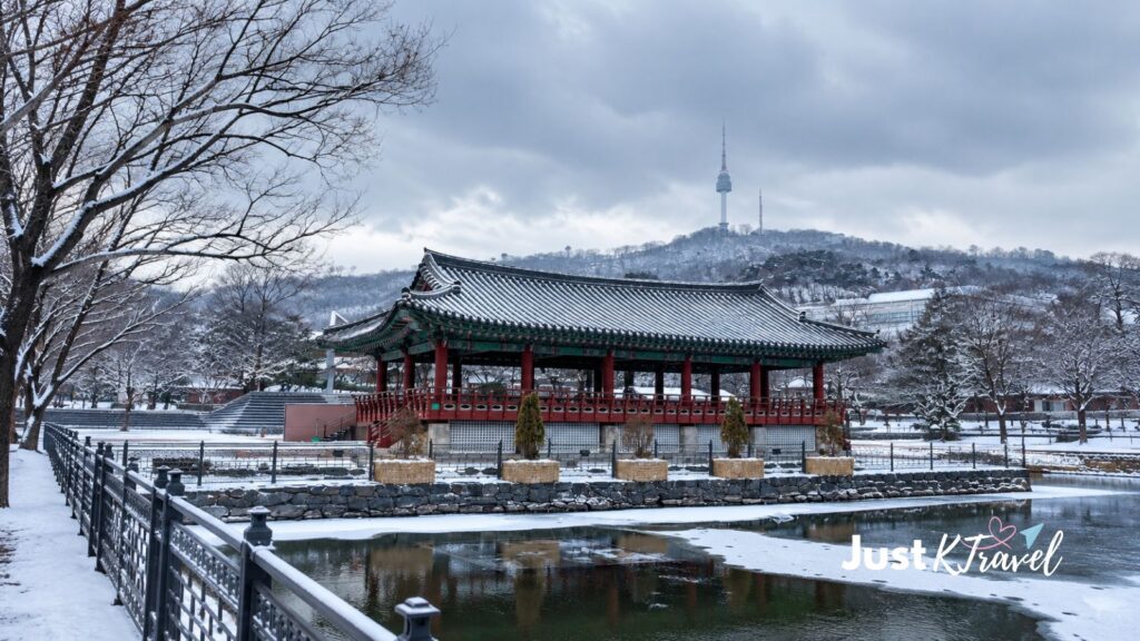 Snow-covered road and traditional hanok pavilion at Namsangol Hanok Village with N Seoul Tower in the background.